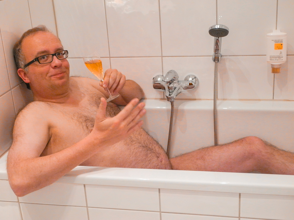 Male nude lies in hotel bathtub enjoying a glass of wine
